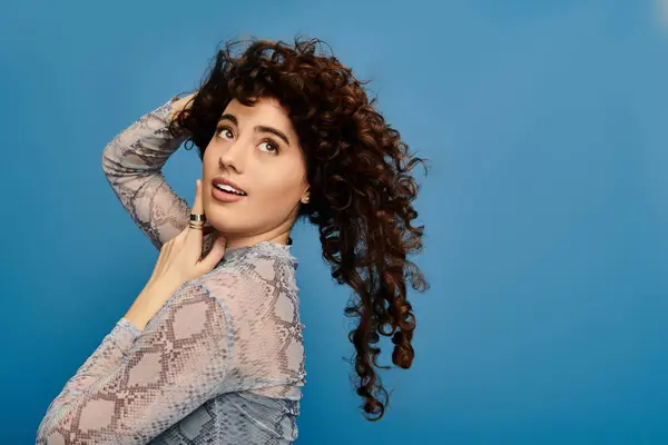 Femme émotionnelle aux cheveux bouclés pose ludique par un mur bleu, capturant un moment élégant. — Photo de stock