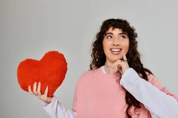 Emotional young woman wearing trendy clothes holds a heart pillow and reflects with joy indoors. — Stock Photo