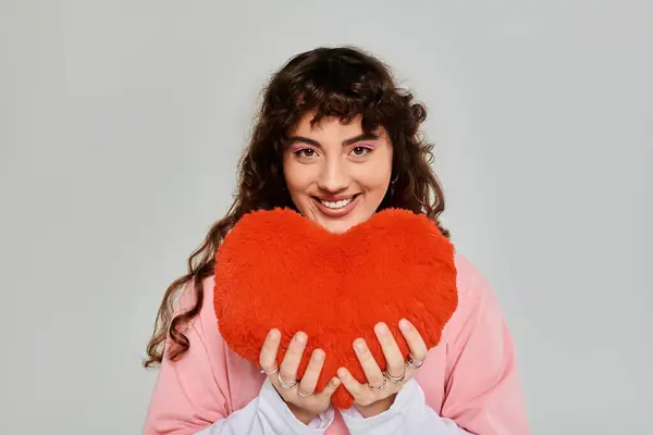Une femme à la mode aux cheveux bouclés tient joyeusement un oreiller en forme de coeur rouge moelleux, exsudant le bonheur. — Photo de stock