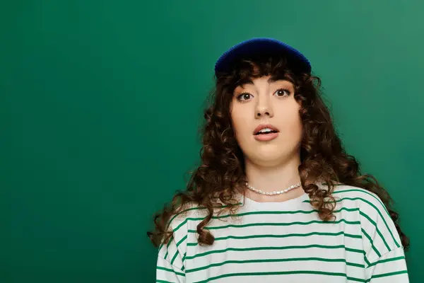 Una mujer joven con el pelo rizado muestra sorpresa mientras usa un sombrero de moda y una camisa a rayas. - foto de stock
