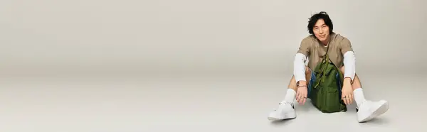 A stylish young Asian man sits on the floor with a trendy backpack, showcasing modern fashion. — Stock Photo