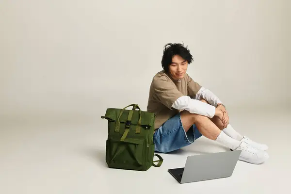 Young man with a trendy outfit smiles while sitting with his laptop and backpack on the floor. — Stock Photo