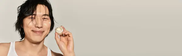 A fashionable young asian man smiles confidently while holding a skincare product in hand. — Stock Photo