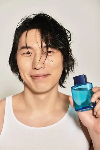 Handsome young asian man smiles while showcasing a trendy fragrance against a minimalist backdrop. — Stock Photo