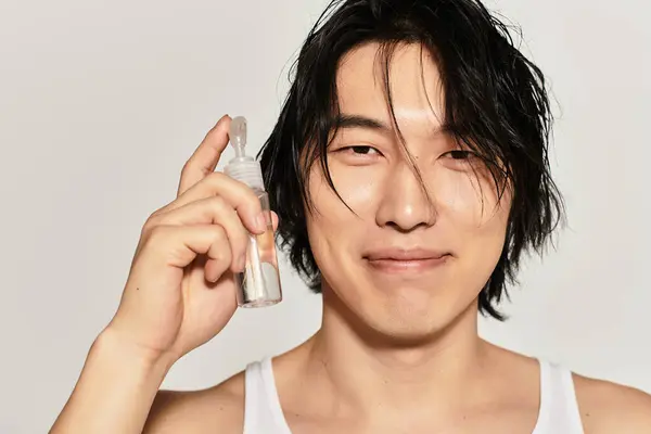 Schöner junger asiatischer Mann mit zerzausten Haaren zeigt lächelnd ein modisches Hautpflegeprodukt. — Stockfoto