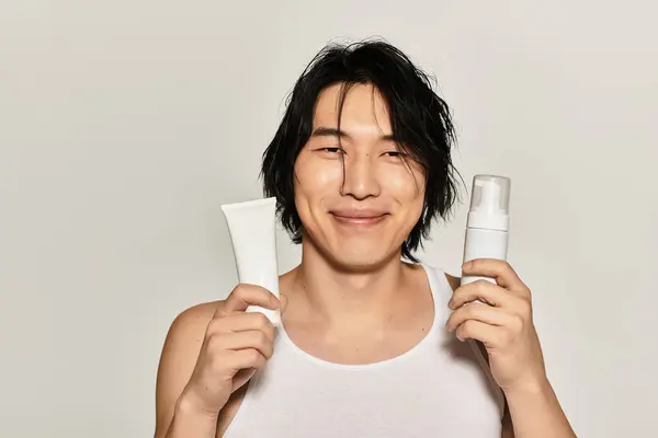 Handsome young man smiling while holding skincare products, radiating modern confidence and style. — Stock Photo