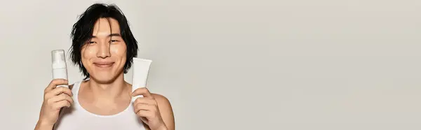 Stylish young Asian man smiles, showcasing trendy skincare products in a modern setting. — Stock Photo