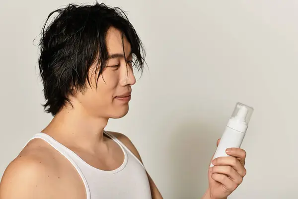 Handsome young asian man with stylish hair smiles while holding a skincare product at home. — Stock Photo