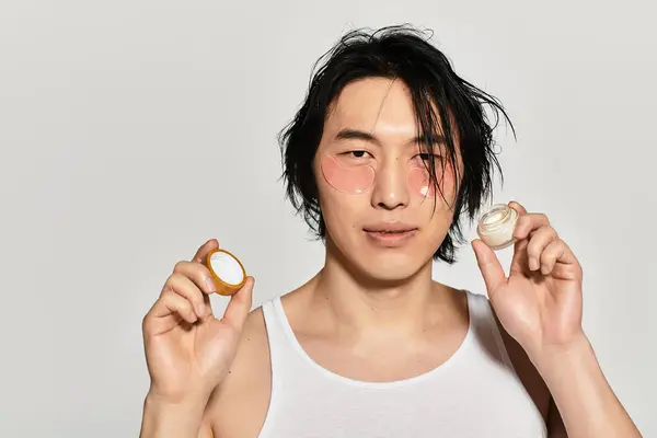 Young asian man holds skincare products with a trendy and appealing look, radiating confidence. — Stock Photo