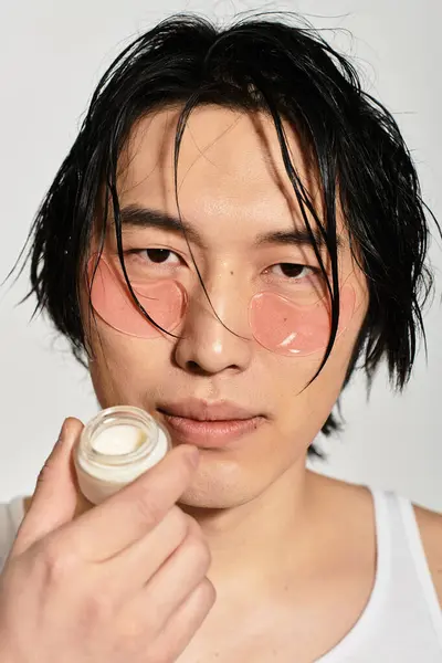 Un joven guapo sostiene un producto para el cuidado de la piel mientras usa gafas de sol rosas redondas de moda. - foto de stock