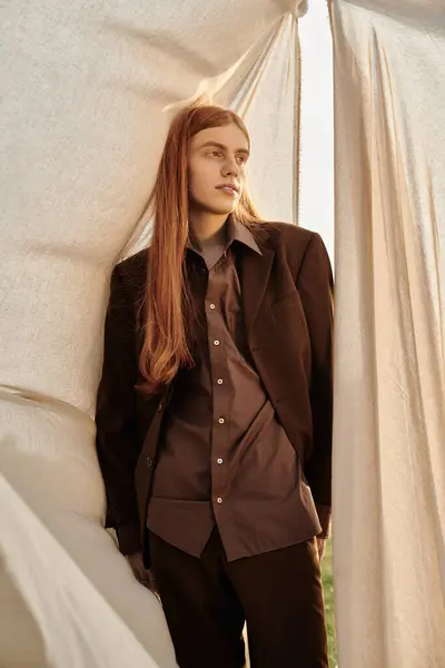 A stylish young man with long hair leans against draped fabric. — Stock Photo