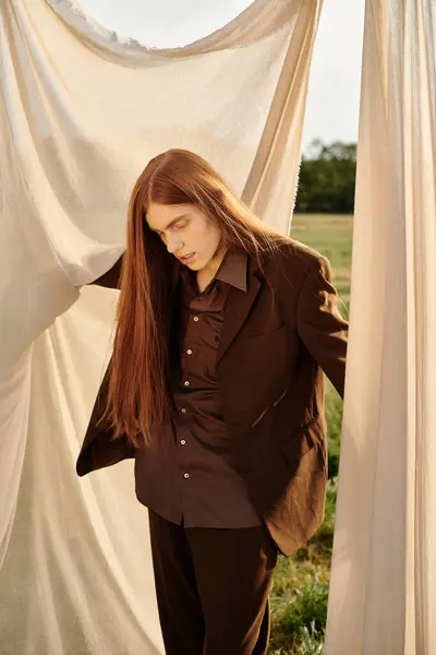 A young, handsome man with long hair stands amidst flowing fabric in a serene outdoors. — Stock Photo