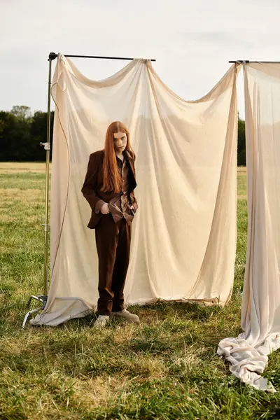 A stylish young man with long hair exudes elegance outdoors by draped fabric. — Stock Photo