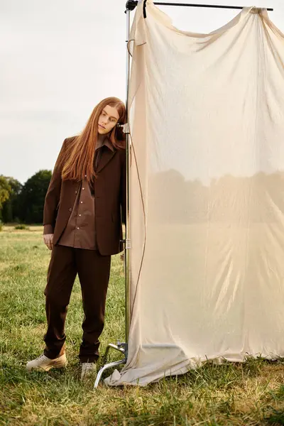 A young man with long hair leans against a fabric backdrop in a peaceful, grassy landscape. — Stock Photo