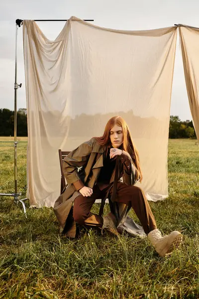 A young man with long hair showcases fashionable attire while seated on a chair in an open field. — Stock Photo