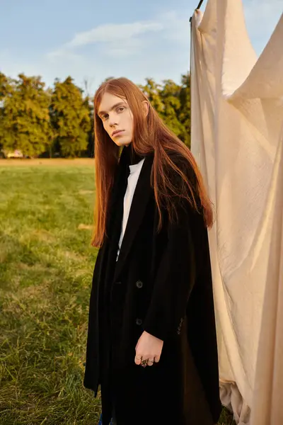 A young man with long hair models stylish clothing in a sunny grassy field. — Stock Photo