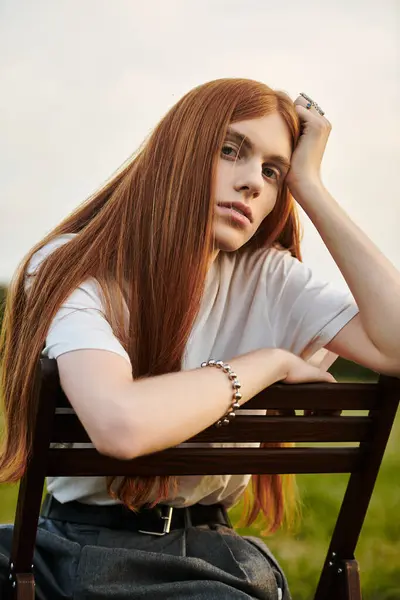 A fashionable young man with long red hair leans thoughtfully on a chair, surrounded by nature. — Stock Photo