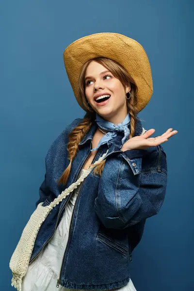 Joyful young woman in a denim jacket and hat against a blue backdrop, embodying spring vibes. — Stock Photo