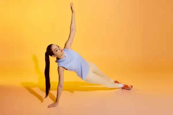 A young woman demonstrates her strength with a side plank on a bright yellow backdrop. — Stock Photo