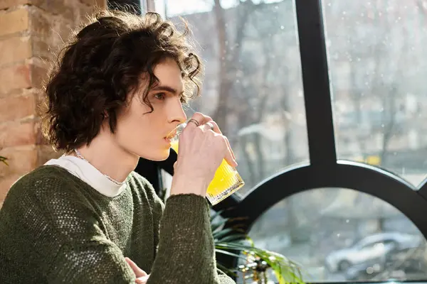 Handsome young guy sips a bright beverage, deep in thought at a charming cafes window. — Stock Photo