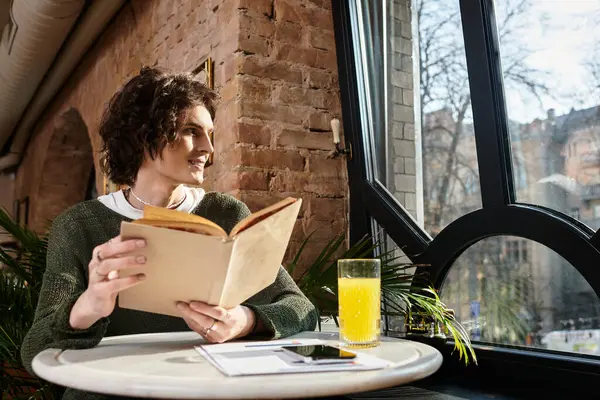 A young man smiles, lost in a book with a drink at a charming cafe. — Stock Photo