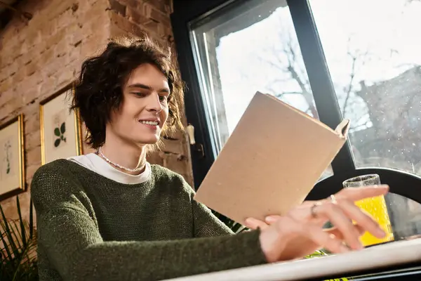 Joven en un café relajándose con un libro y una bebida refrescante mientras entra la luz del sol. - foto de stock