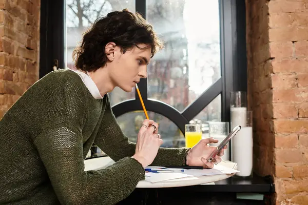 Le jeune homme est assis dans un café, profondément dans la pensée tout en tenant un crayon dans sa bouche et son téléphone. — Photo de stock