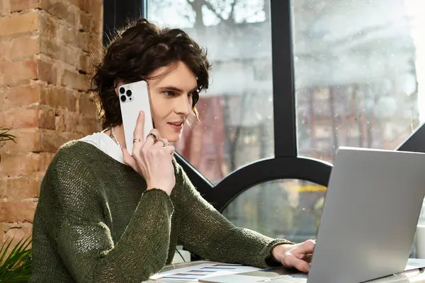 At a cozy cafe, a young man chats on the phone while working on his laptop. — Stock Photo