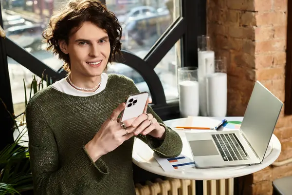 A handsome young man lounges in a cafe, focused on his smartphone amid paperwork and a laptop. — Stock Photo