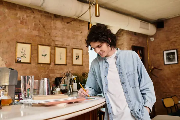 A young man with stylish hair sits at a cafe, happily drawing with a pencil while sipping a drink. — Stock Photo