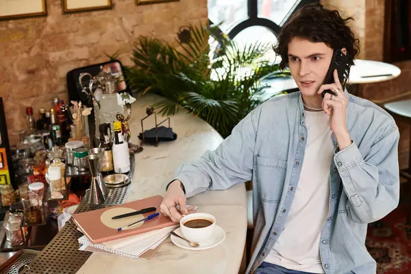 A young man with a stylish look sips coffee while engaged in a phone conversation at a cozy cafe. — Stock Photo
