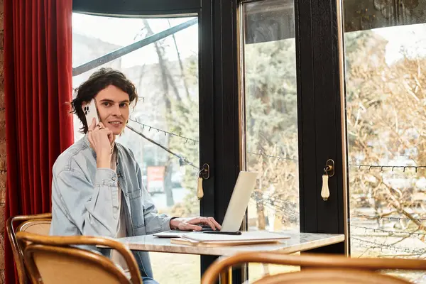 Handsome young guy sits at a cafe table, talking on the phone and working on his laptop, relaxed. — Stock Photo