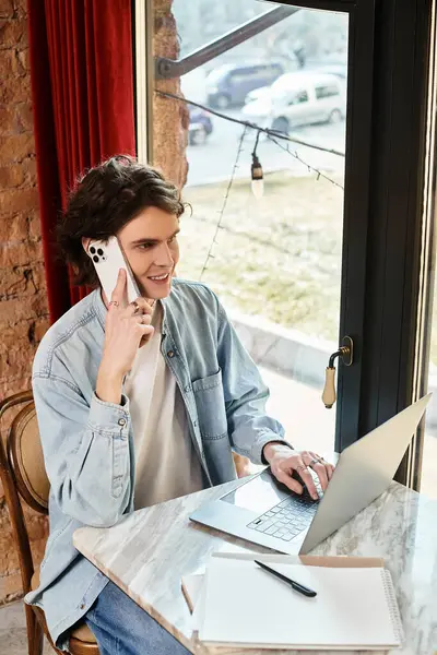 Young man sits at a stylish cafe, chatting on the phone and working on his laptop, fully absorbed. — Stock Photo
