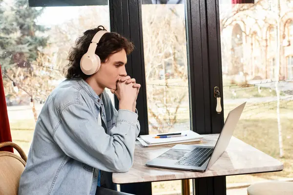 Un jeune beau garçon s'assoit soigneusement dans un café, concentré sur son ordinateur portable avec des écouteurs sur. — Photo de stock