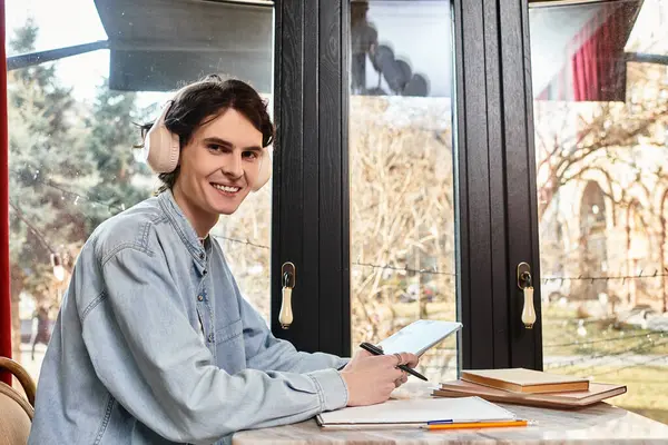 Cheerful young man with headphones sits at a table, studying and enjoying a calm cafe atmosphere. — Stock Photo