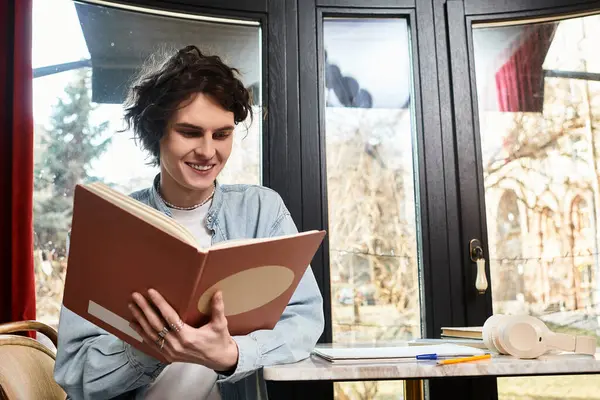 Um jovem e bonito homem se senta em uma mesa de café, profundamente imerso na leitura de um livro com um sorriso. — Fotografia de Stock