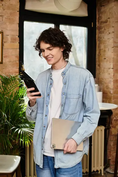 A young, attractive guy stands in a cafe, holding a smartphone and a laptop, relaxed and happy. — Stock Photo