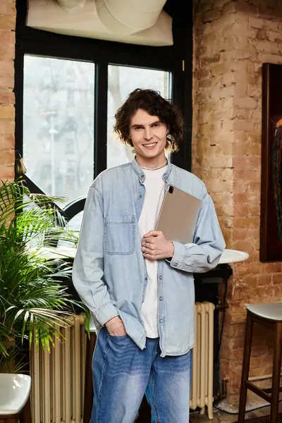 A confident young man stands in a vibrant cafe, holding a laptop and enjoying the vibe. — Stock Photo