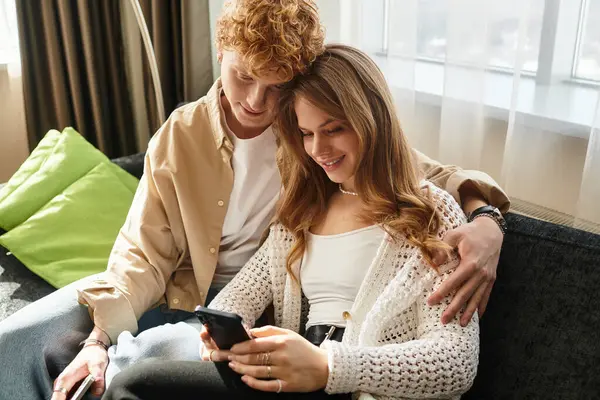 A loving couple cuddles on a sofa, sharing laughter with a mobile device in their hotel room. — Stock Photo