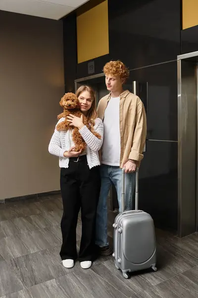 Una joven pareja se encuentra en un moderno vestíbulo del hotel, abrazando a su adorable cachorro con alegría. - foto de stock