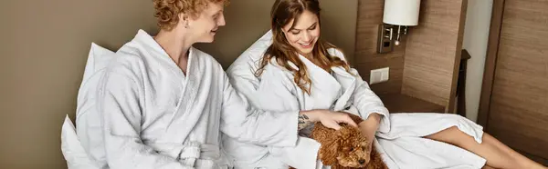 Una giovane coppia in accappatoi accoglienti si rilassa in una camera d'albergo, condividendo momenti teneri con il loro cane. — Foto stock
