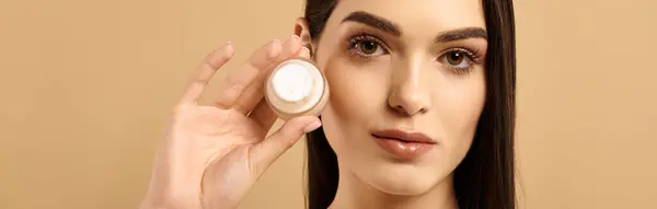 A young woman displays her radiant skin while holding a skin care product in a studio, banner — Stock Photo