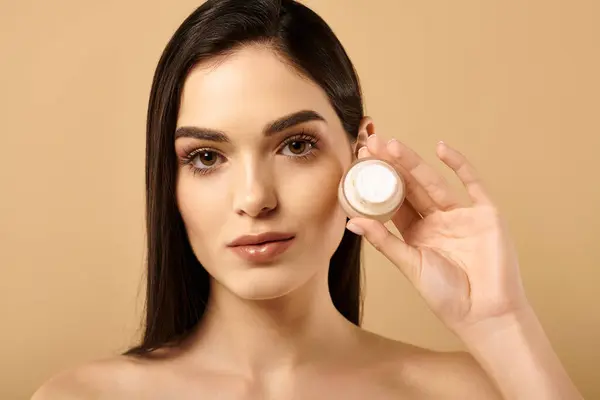 A young woman showcases her healthy, radiant skin while holding a beauty product in a studio. — Stock Photo