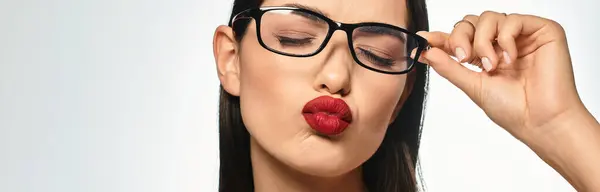 A young woman with brown hair and red lips playfully adjusts her glasses on a grey backdrop, banner — Stock Photo