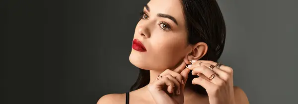 A beautiful young woman with brown hair and red lips gracefully adorns herself with elegant jewelry. — Stock Photo