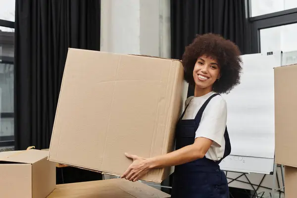 A cheerful woman lifts a big cardboard box in a modern space, packing their belongings. — Stock Photo