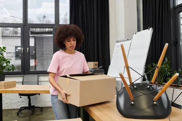 A young woman packs a box amid furniture and moving supplies in a modern office. — Stock Photo