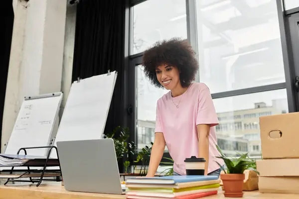 Una mujer con una camisa rosa claro se centra en su computadora portátil, rodeada de cajas de embalaje y plantas. - foto de stock