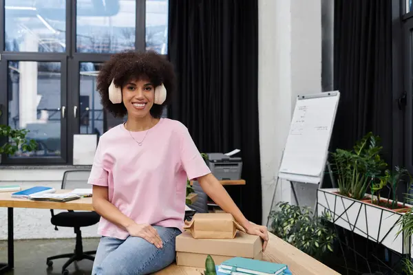 A young woman with curly hair wearing headphones stands next to packed boxes in a vibrant office. — Stock Photo