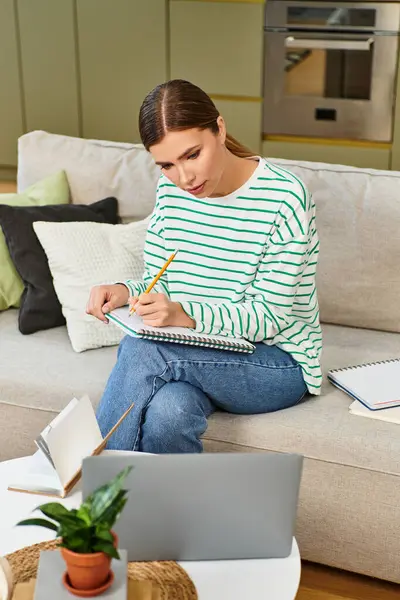 Beautiful woman sits on a couch in a modern living room, focused on taking notes while studying. — Stock Photo